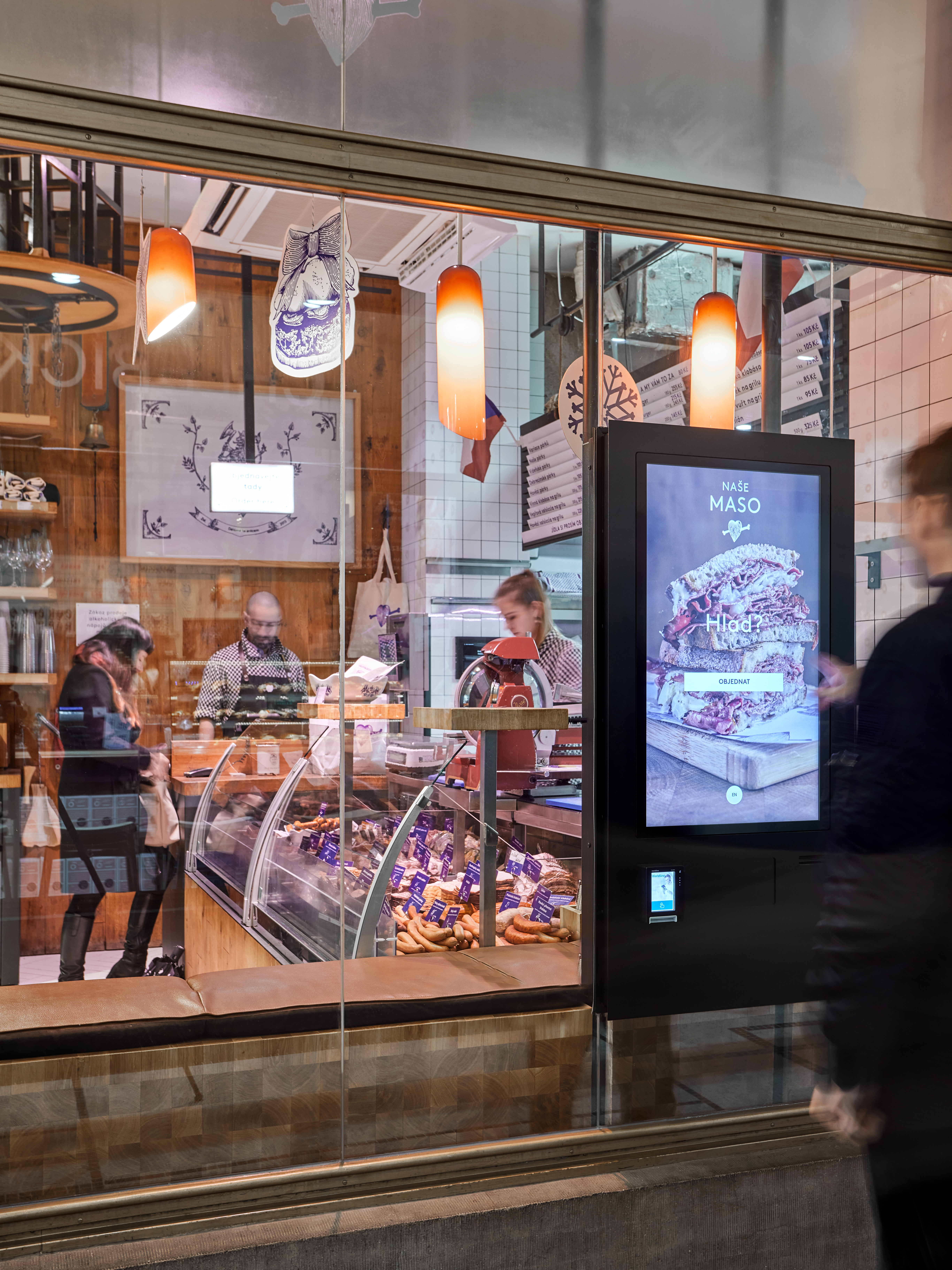 A compact self-service kiosk counter solution in a bright corporate reception area, featuring a wide touchscreen flush-mounted into a brushed aluminum housing, with an integrated POS card reader and contactless symbol, plus a small built-in receipt slot below. The kiosk sits on a light oak reception desk with a minimalist arrangement of folders and a monitor stand in the background, intentionally blurred. Soft, diffused daylight enters from large windows to the left, mixing with subtle recessed ceiling lights to create a professional, welcoming atmosphere. Captured from a slightly elevated three-quarter angle with sharp focus on the kiosk, the photographic image highlights the unified kiosk, point-of-sale, and payment terminal integration in a clean, businesslike setting.